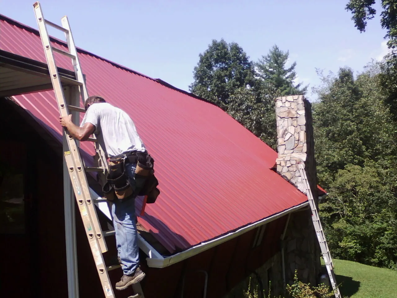 Roofer on a ladder beside a red metal roof and stone chimney — Soffit & Fascia Repair by Roofing Brothers in Sodus Point